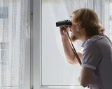 Uomo con binocolo spia dalla finestra, forse per atti persecutori vicino di casa.