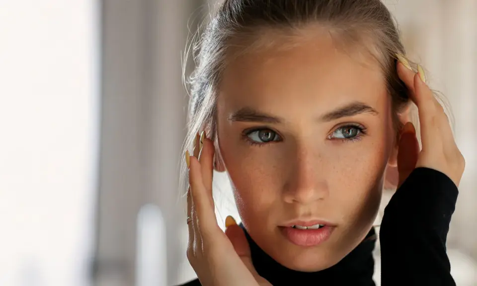 Primo piano di una giovane donna con lentiggini, che si tocca il viso. Il suo sguardo intenso e le sopracciglia leggermente alzate suggeriscono sorpresa o curiosità, rivelando il significato psicologico di questo gesto.