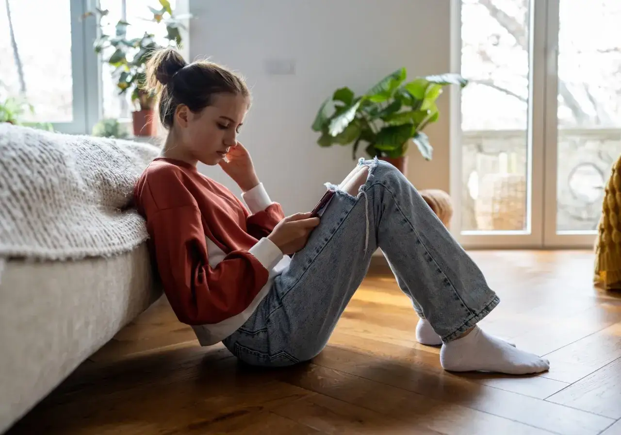 Ragazza pensierosa seduta sul pavimento, con jeans strappati e felpa arancione. Forse sta pensando di andare via di casa per colpa dei genitori.
