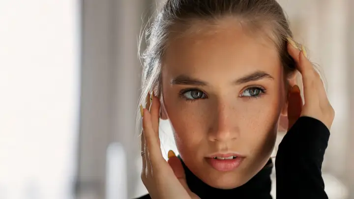 Primo piano di una giovane donna con lentiggini, che si tocca il viso. Il suo sguardo intenso e le sopracciglia leggermente alzate suggeriscono sorpresa o curiosità, rivelando il significato psicologico di questo gesto.